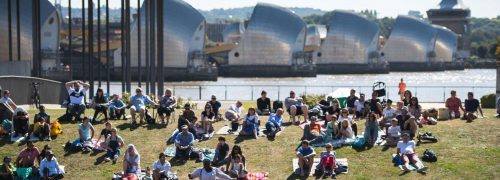 Image shows people sitting on the grass at Thames Barrier Park, enjoying an event.