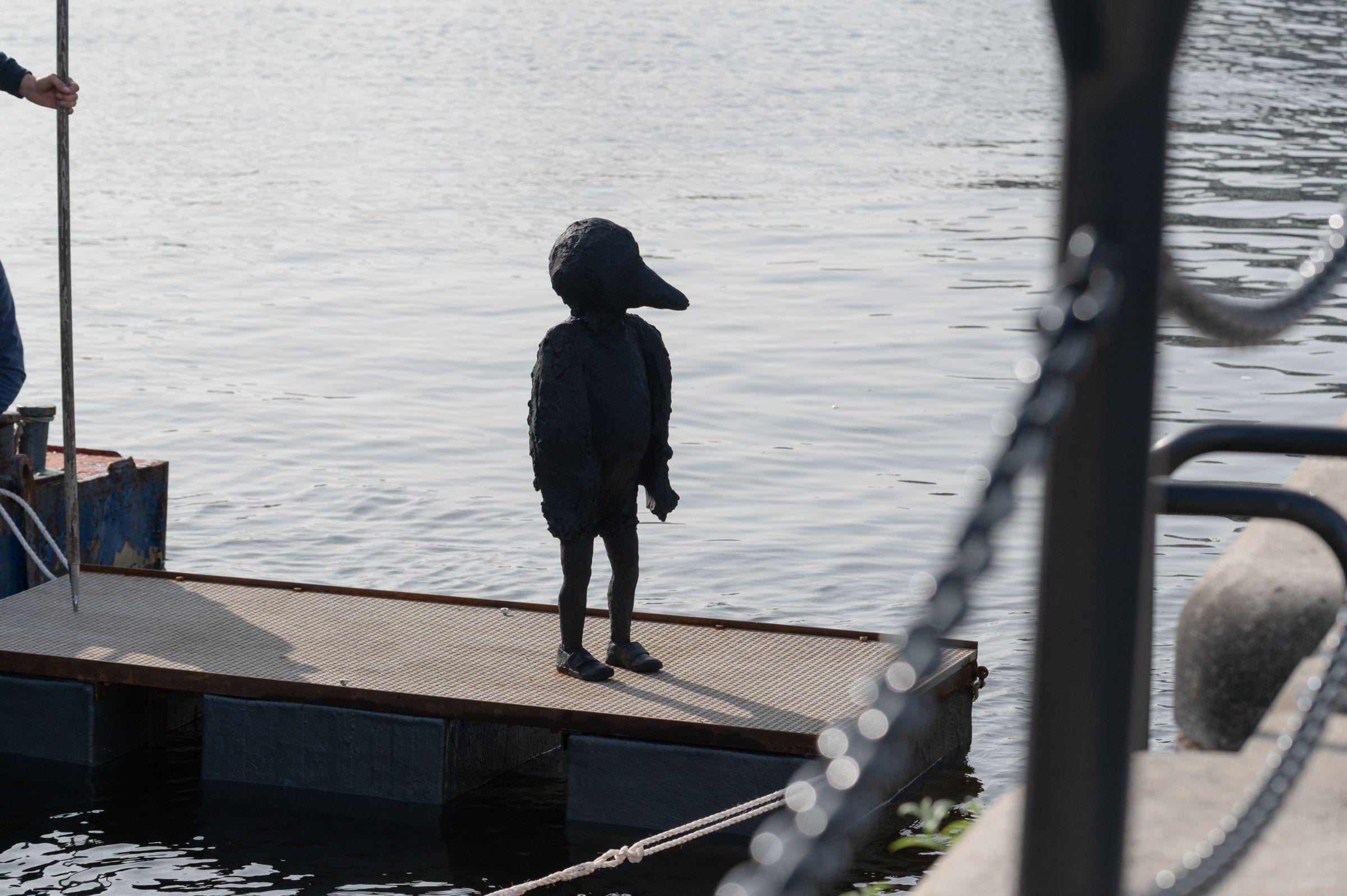 Statue that looks like a child with a bird's head, floating on a platform in the dock