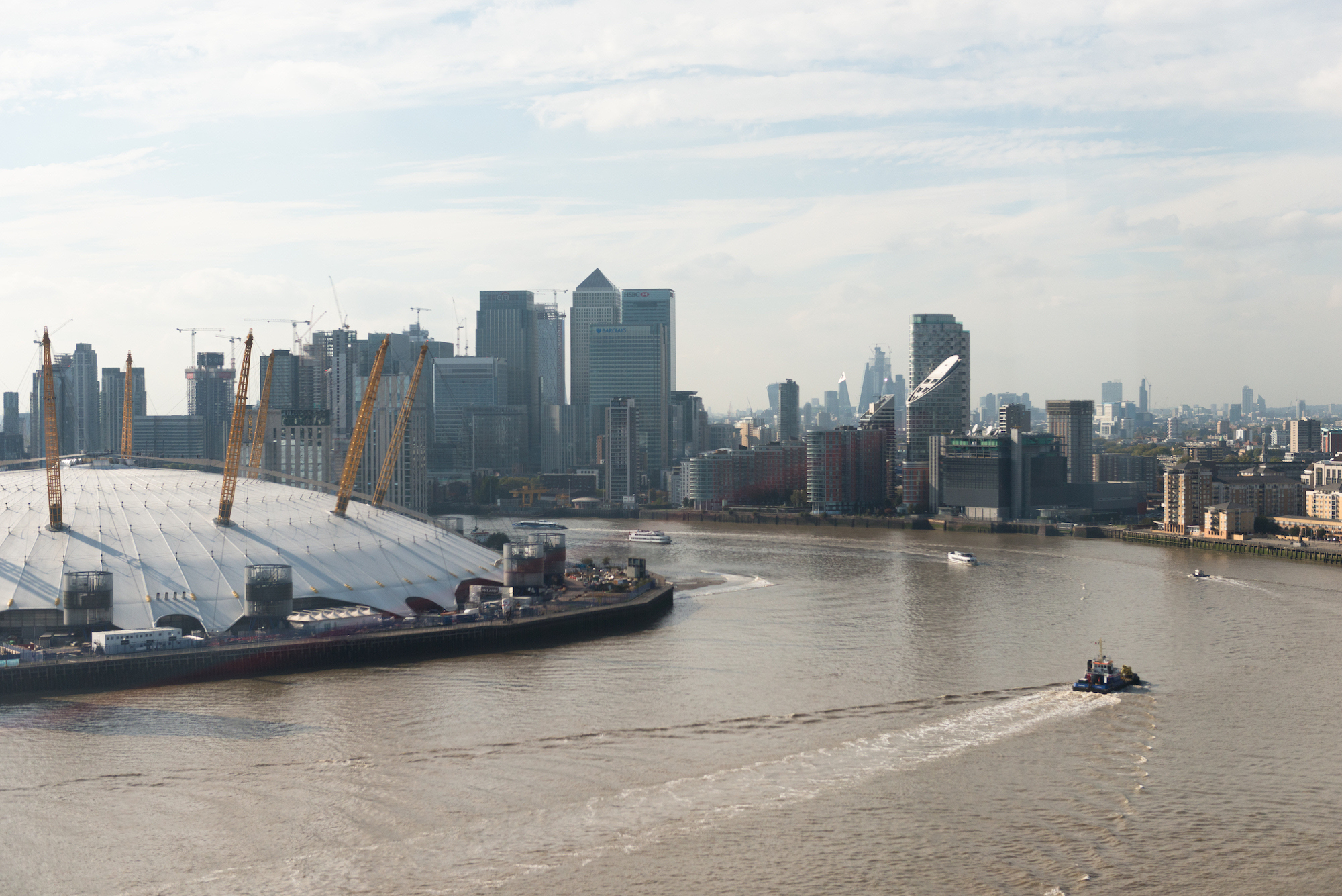 the millennium dome with canary wharf in the background, and the wide brown river thames with a boat going by