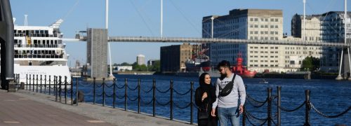 Two people walking along the Royal Docks with Millennium Mills in the background