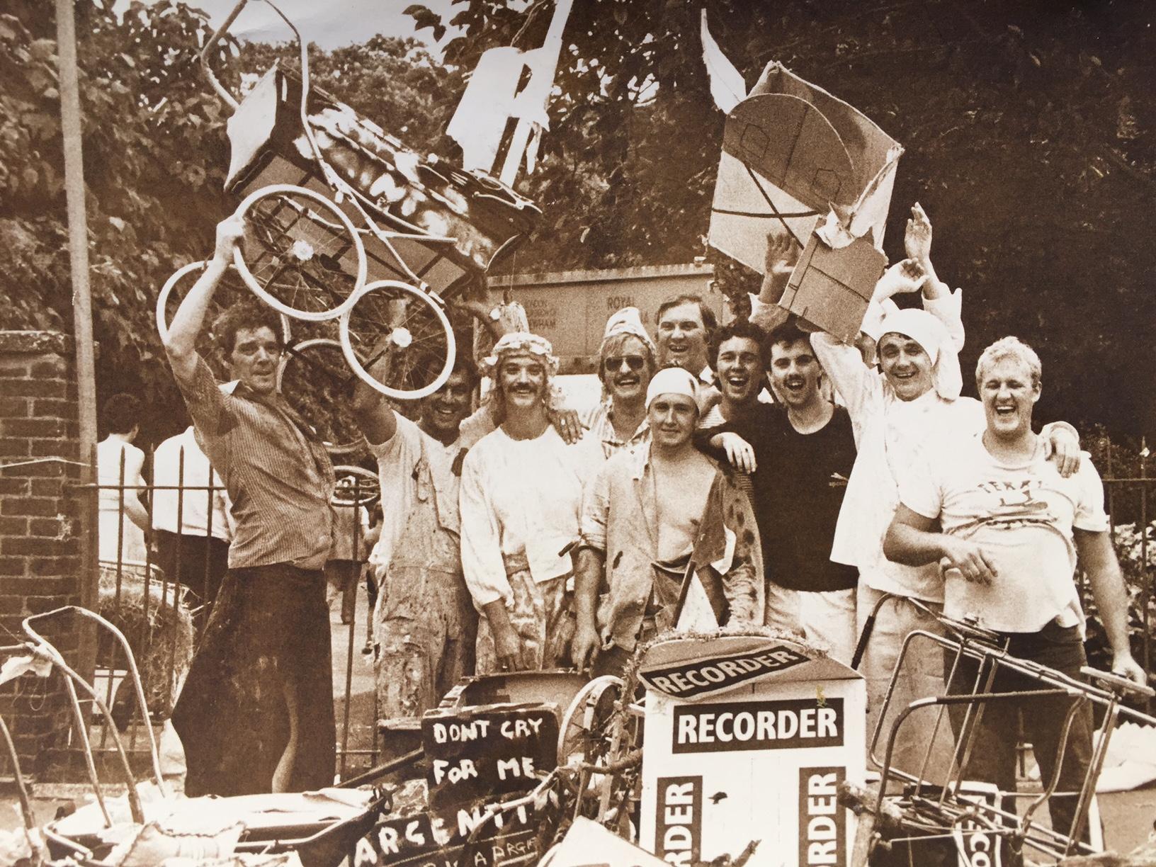 Group of men smiling, wearing costumes. One is waving a pram in the air.