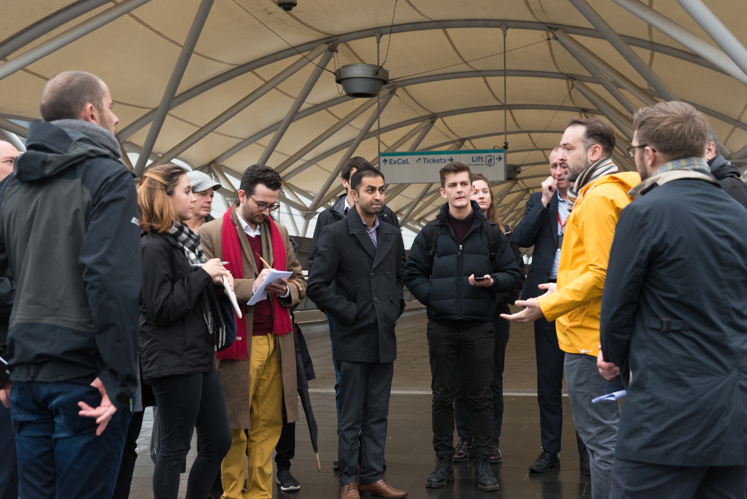 Group of people standing in the walkway to the ExCeL centre listening to a man speaking