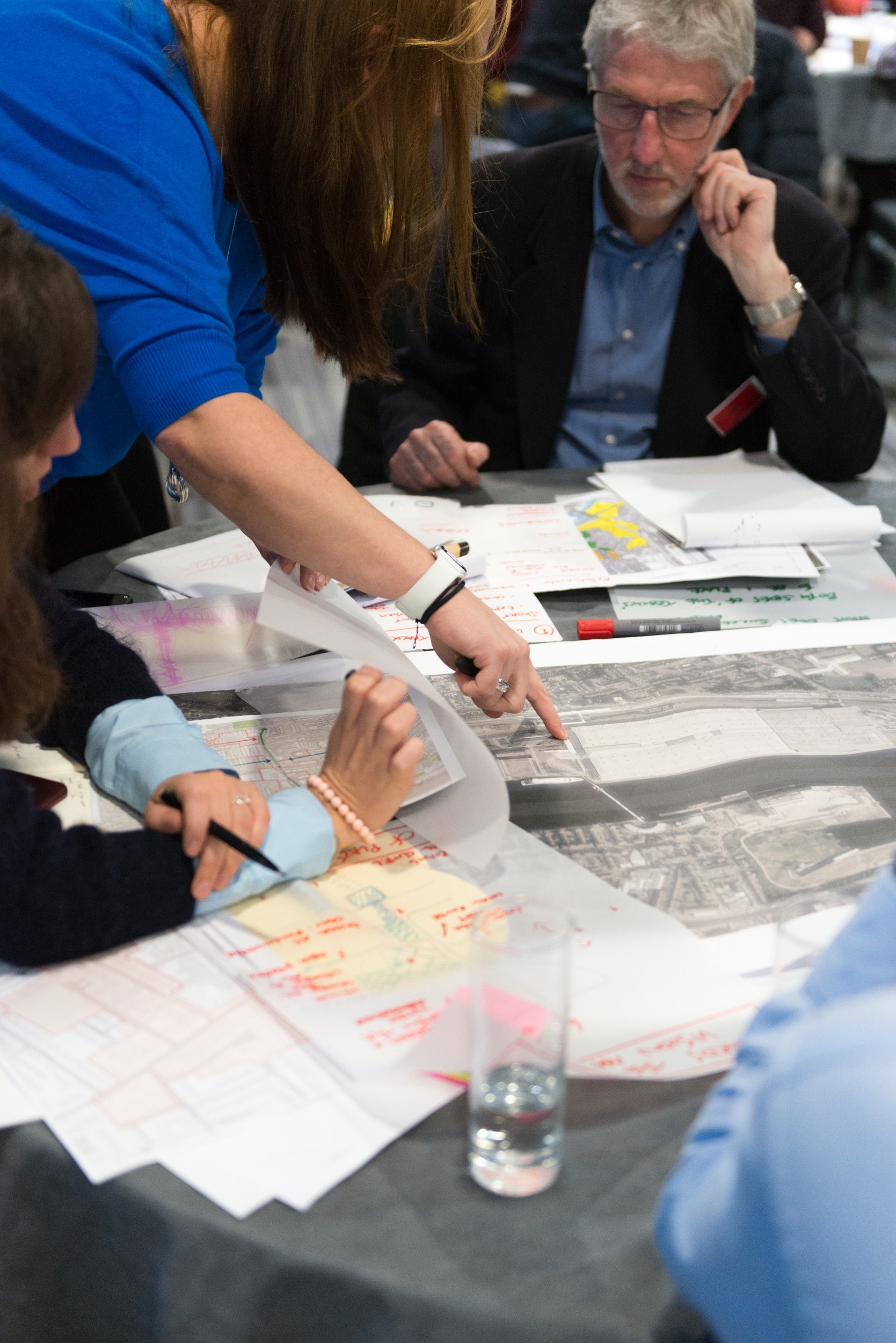Hand pointing at a pile of maps and diagrams, with people sitting around a table