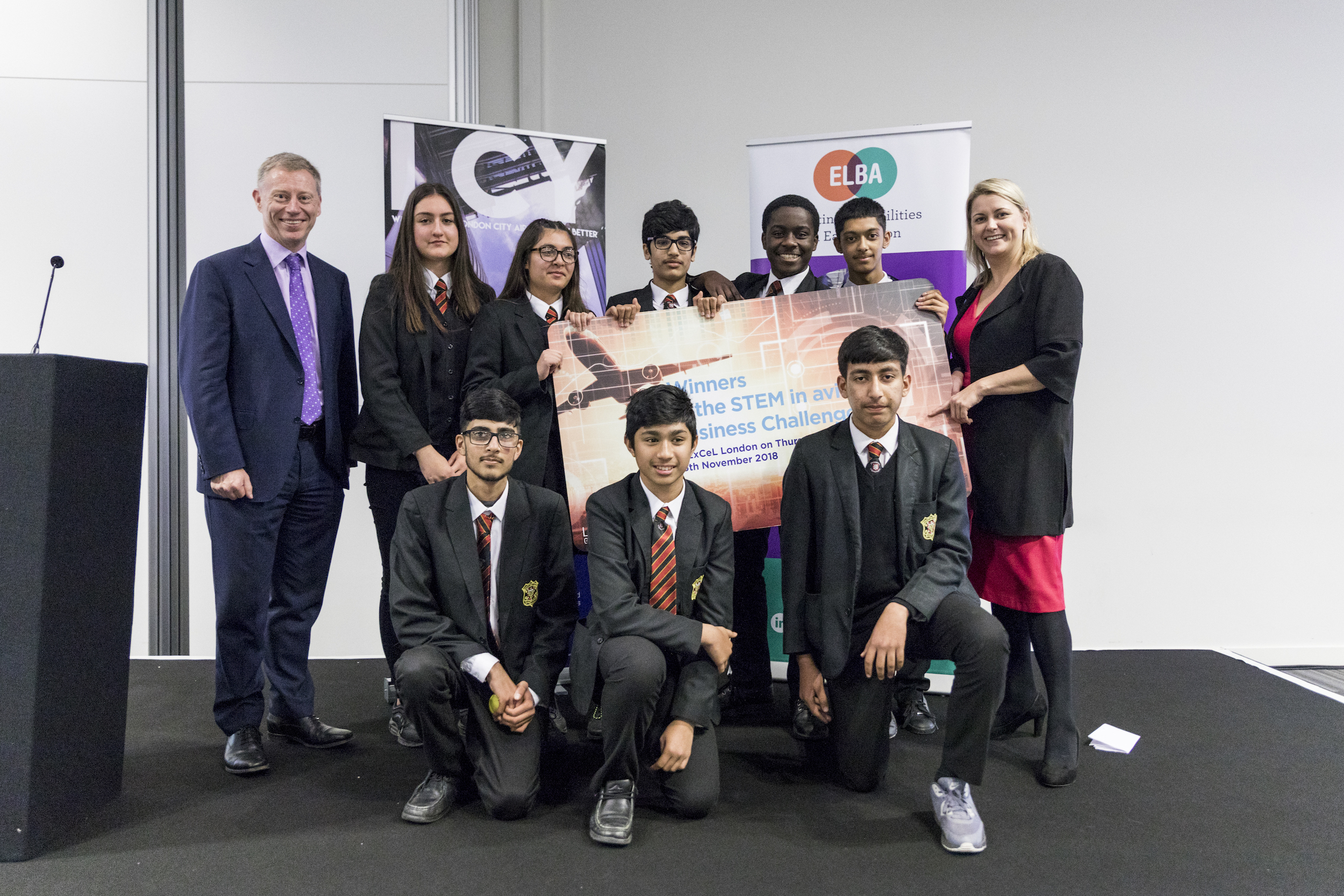 The winning team holding a plaque on stage, flanked by Robert Sinclair and Baroness Sugg