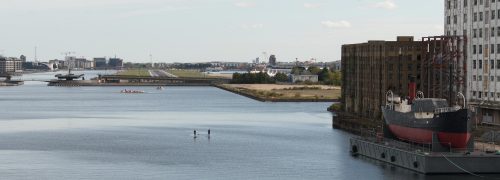 Looking down Royal Victoria Dock with paddleboarders and historic ship and buildings to the right